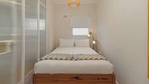 Un lit bien fait, avec une couvre-lit à carreaux et des serviettes pliées, une tête de lit en bois, un luminaire suspendu en fibres tressées et une fenêtre avec des stores.