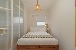 Ein ordentlich gemachtes Bett mit karierter Bettdecke und zusammengerollten Handtüchern, ein Holz-Kopfbrett, eine hängende Webdekorleuchte und ein Fenster mit Jalousien.