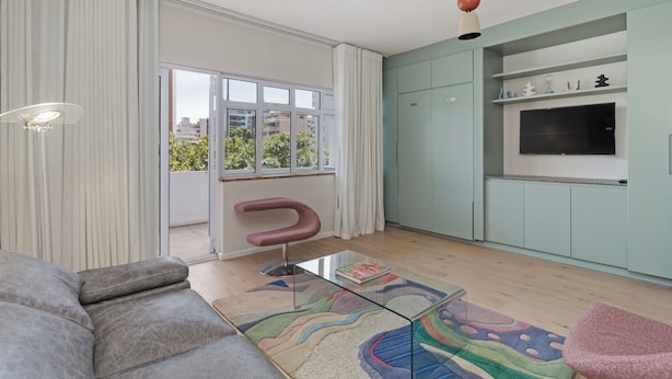 Un salon moderne avec un canapé, une table basse en verre, un tapis coloré, un fauteuil rose et un téléviseur fixé sur un meuble vert clair.