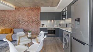 A modern kitchen with a built-in washing machine, a dining area with a glass table and white chairs, and a brick wall. - Stylish Stay at The Signature | Central and Modern (Cape Town)