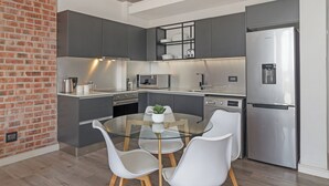 A modern kitchen with grey cabinets, a stainless steel refrigerator, a glass dining table with white chairs, and a brick wall. - Stylish Stay at The Signature | Central and Modern (Cape Town)