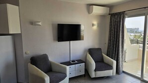 Ett modernt hotellrum med en platt-TV som är monterad på väggen, två fåtöljer, ett litet sidobord och ett stort fönster med gardiner.
