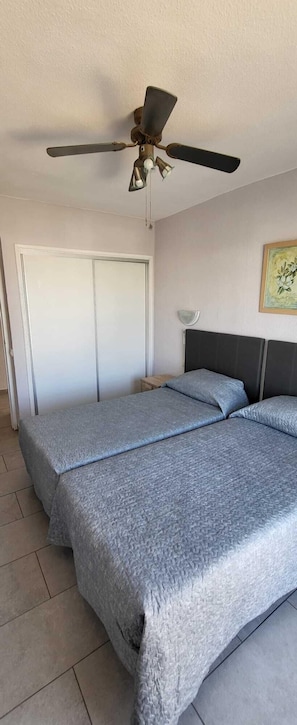 A bedroom with two beds, a ceiling fan, a door, and a wall-mounted light fixture. - Santa Maria 144 - Two Bed (Costa Adeje)