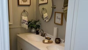 Un baño con tocador de doble lavabo, un espejo, un perchero con toalla, un cuadro enmarco y una lámpara de pared.