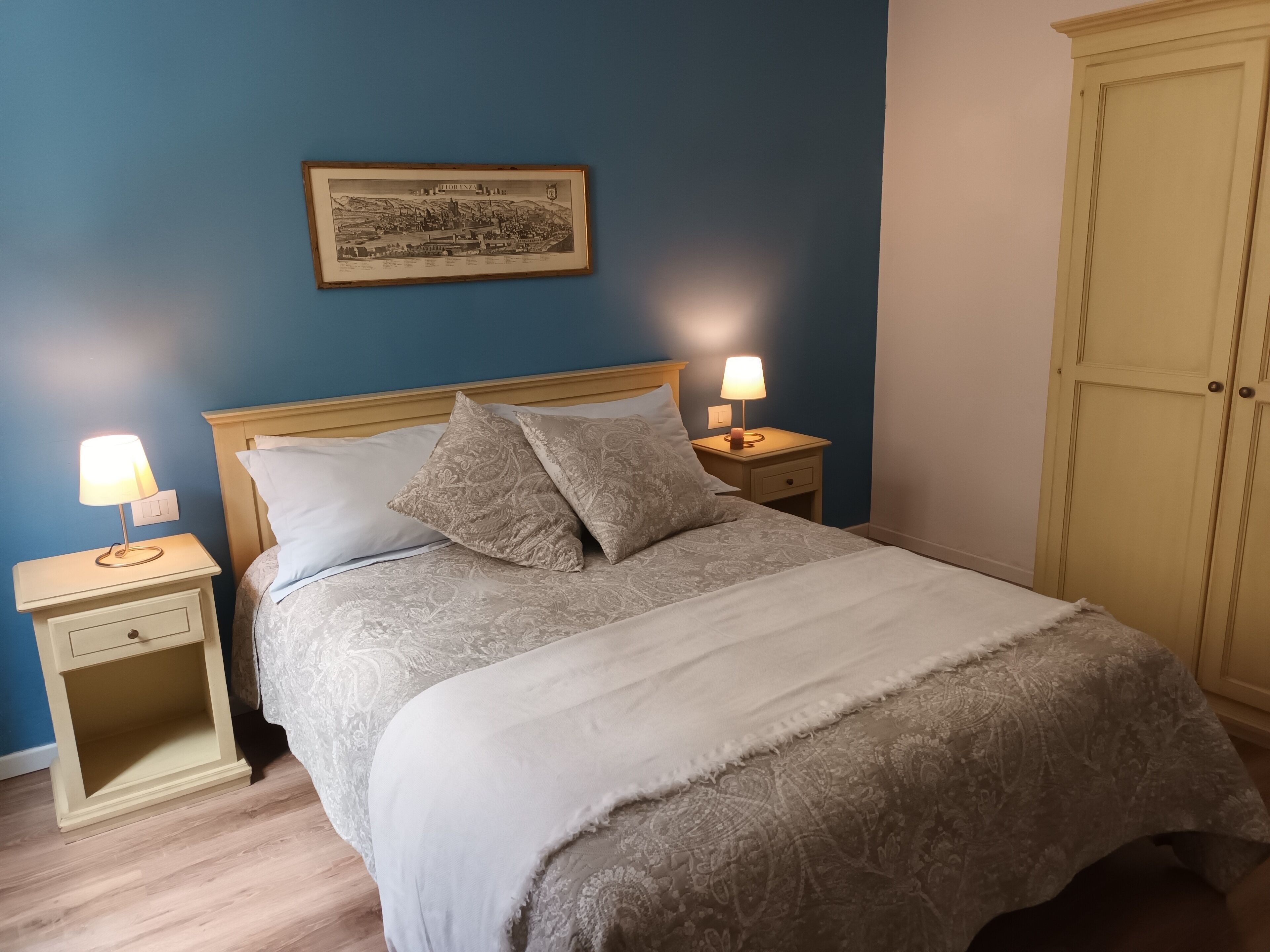 A bedroom with a bed, two bedside tables with lamps, a nightstand drawer, a framed picture on the wall, and a wardrobe.