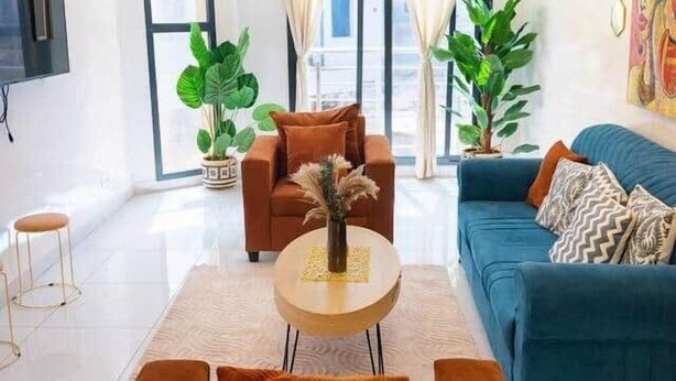 A modern living room with a sofa, armchairs, and a coffee table. There are large windows with curtains, a flat-screen TV mounted on the wall, and a painting on the wall. - Bright Designer 2BR/3BA | Balcony | Washer | Almadies (Dakar)