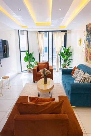 Ein modernes Wohnzimmer mit einer Couch, Sesseln und einem Couchtisch. Große Fenster mit Vorhängen, ein wandmontierter Flachbildfernseher und ein Gemälde an der Wand.