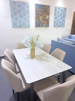Un comedor con una mesa blanca, sillas color beige y un jarrón con flores. Hay tres obras de arte en la pared.