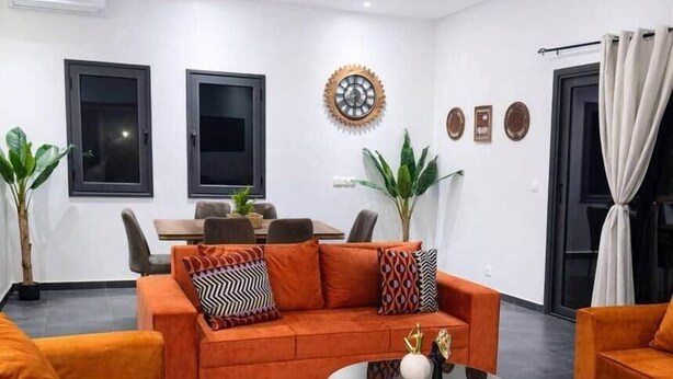 Ein modernes Wohnzimmer mit orangefarbenen Sofas und einem Glastisch. Es gibt einen Essbereich mit Tisch und Stühlen, und eine große Uhr an der Wand.