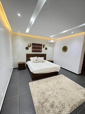 Une chambre à coucher moderne avec un grand lit, une table de chevet, une commode, un tapis à motifs et des œuvres d’art décoratives accrochées au mur.