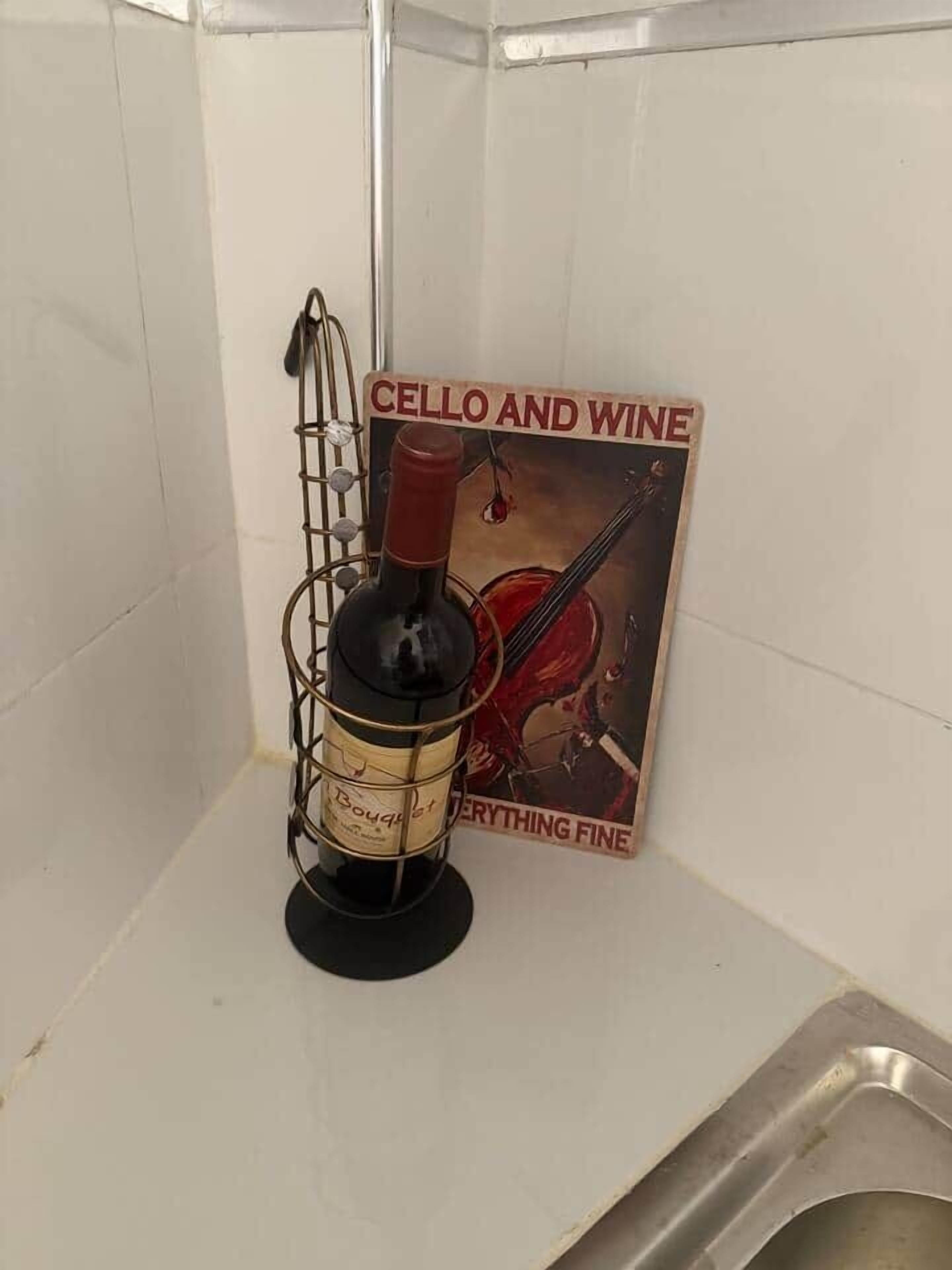 Una botella de vino sobre un soporte metálico con un letrero que dice "CELLO AND WINE" y "EVERYTHING FINE" en una habitación con paredes de azulejos blancos y un lavamanos metálico.