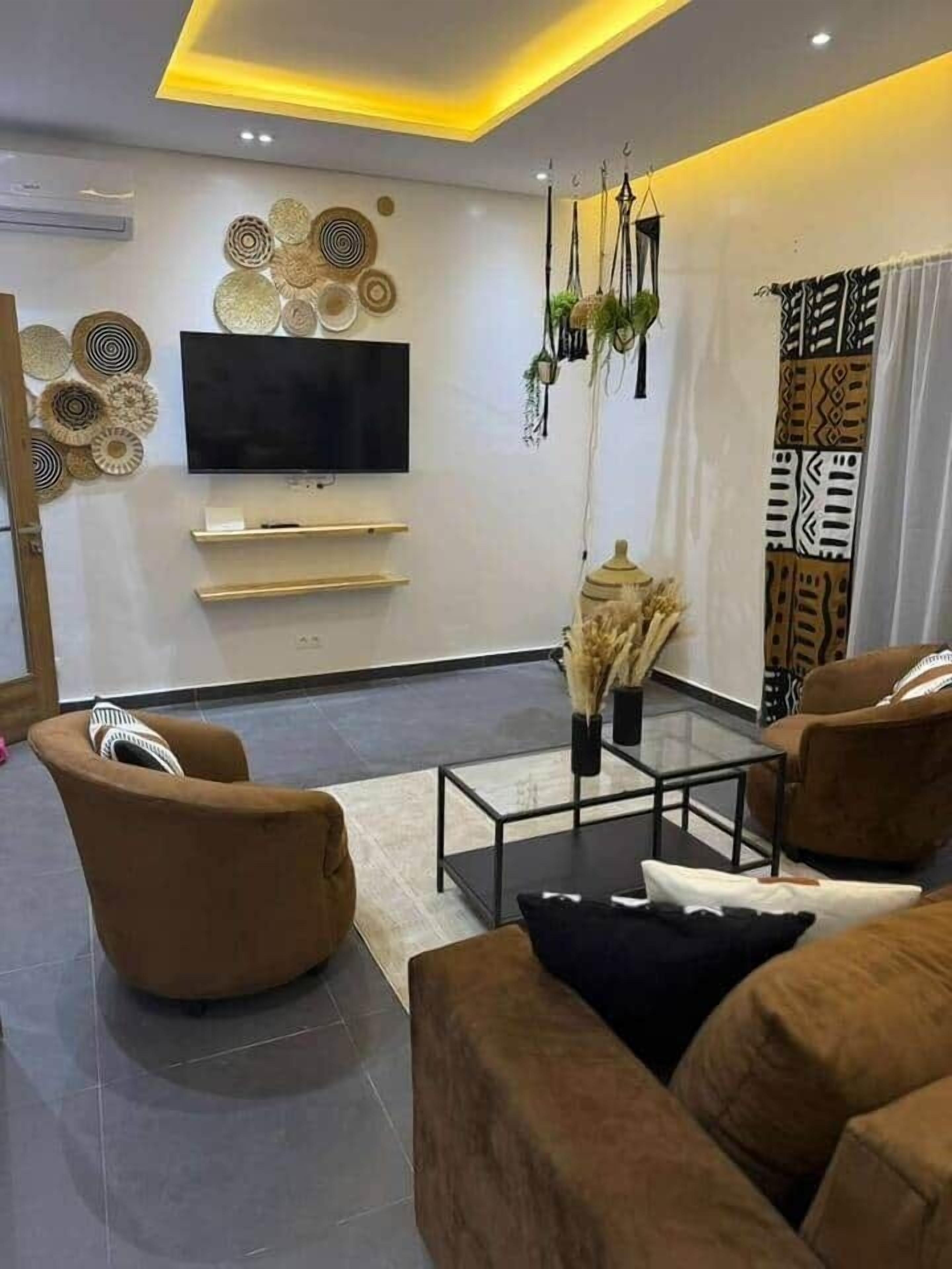 A modern living room with a flat-screen TV, brown sofas, a glass coffee table, decorative wall art, and a patterned curtain.