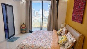 Ein Schlafzimmer mit Bett, Nachttisch, Lampe, Fenster mit Vorhängen und Blick auf Gebäude.