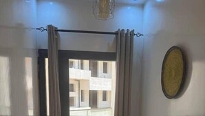 Ein Zimmer mit großem Fenster, Vorhang, Deckenleuchte und einer an der Wand befestigten Dekoration.