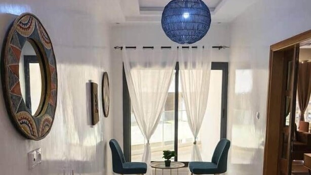 A modern living room with a glass coffee table, two blue chairs, a wooden side table, and a large blue pendant light. - Spacious 2BR Almadies • Rooftop • Bright & Breezy (Dakar)
