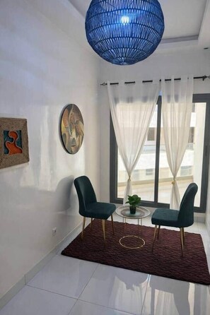 A modern living room with two green chairs, a round coffee table, a blue hanging light fixture, and a view of the outside through large windows with white curtains. - Spacious 2BR Almadies • Rooftop • Bright & Breezy (Dakar)