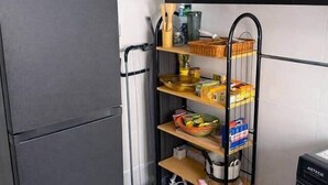 A compact kitchen with a black refrigerator, a metal shelf unit with various food items, a washing machine, and a small stool. - Spacious 2BR Almadies • Rooftop • Bright & Breezy (Dakar)