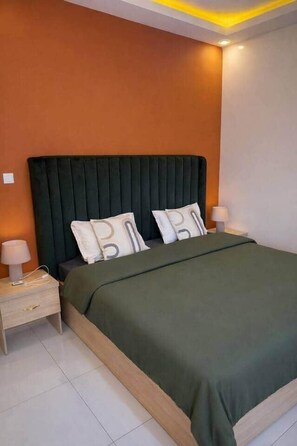 A modern bedroom with a dark green upholstered headboard, a green bedspread, and two white pillows with a gray pattern. There are two bedside tables with lamps and a small drawer unit. - Spacious 2BR Almadies • Rooftop • Bright & Breezy (Dakar)