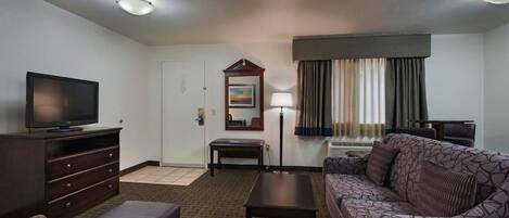 A hotel room with a television, a sofa, a coffee table, a desk, a lamp, a mirror, and a window with curtains.