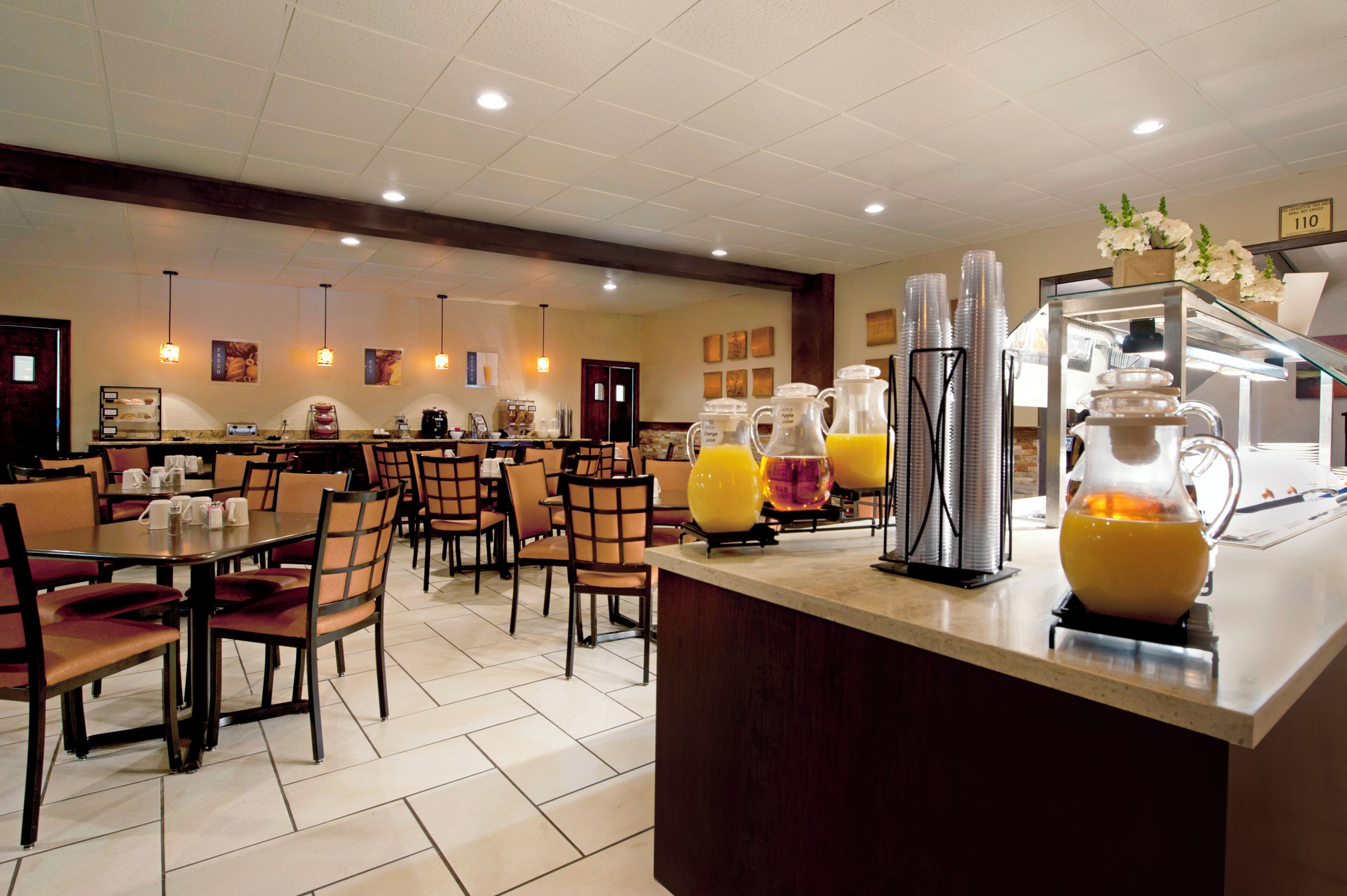A hotel breakfast area with tables, chairs, a juice station, and a refrigerator.