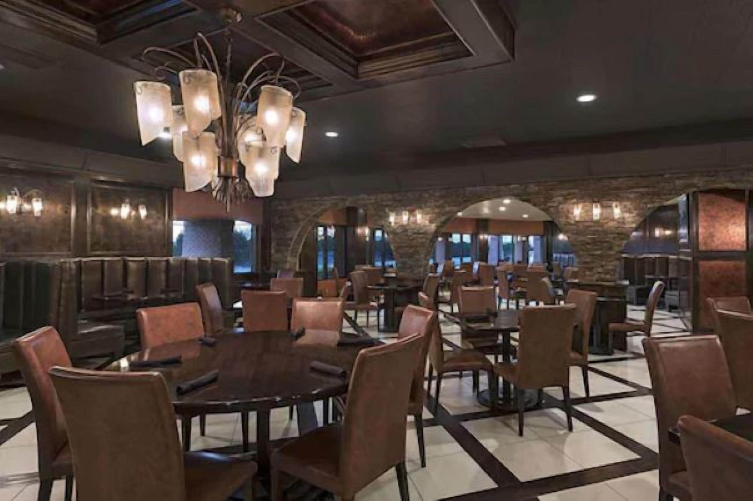 A spacious dining area with multiple tables and chairs, a central chandelier, and stone walls.