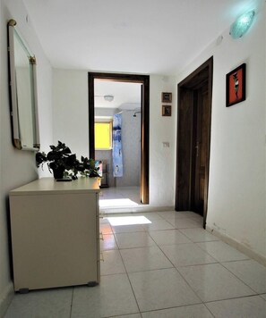 Un pasillo con suelo de baldosas blancas, un escritorio con planta en maceta, un espejo y una puerta que da a otra habitación.