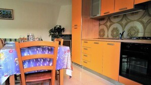 Ruokailutila, jossa on pöytä, jonka päällä on pöytäliina, sekä tuoleja, ja keittiö, jossa on oranssit kaapit ja liesi.