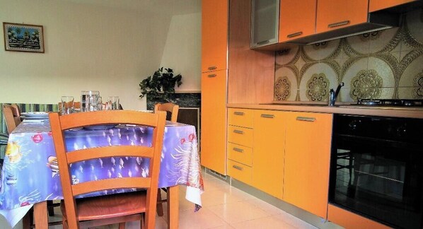 Un comedor con una mesa cubierta por un mantel, sillas y una cocina con armarios naranjas y una estufa.