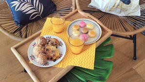 Two wicker chairs with floral cushions, a tray with pastries, a plate of cookies, a glass of orange juice, and a plate of macarons.