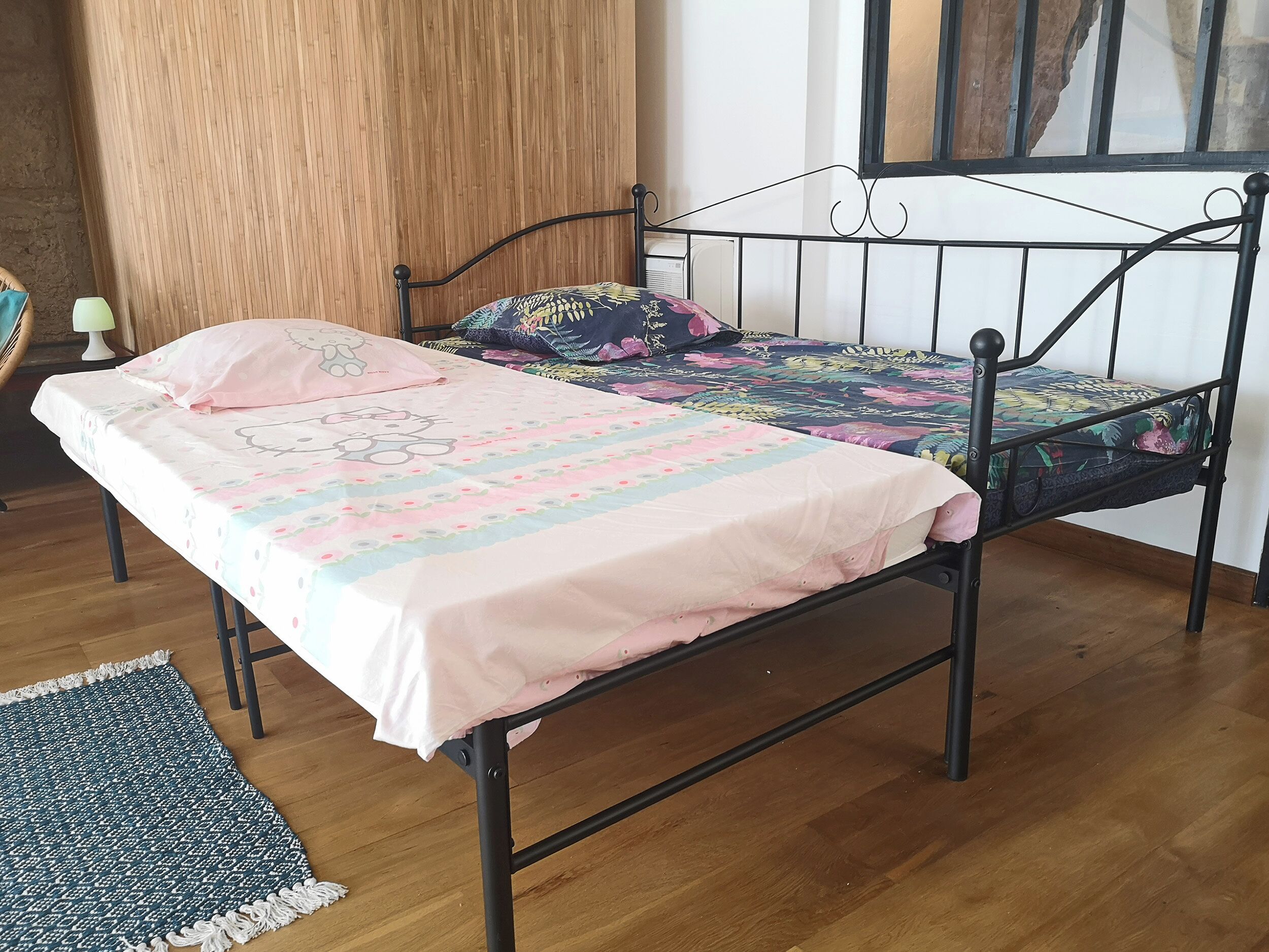 Un lit avec une couvre-lit à motifs floraux et un coussin à l’effigie de Hello Kitty, un plancher en bois et un petit tapis.