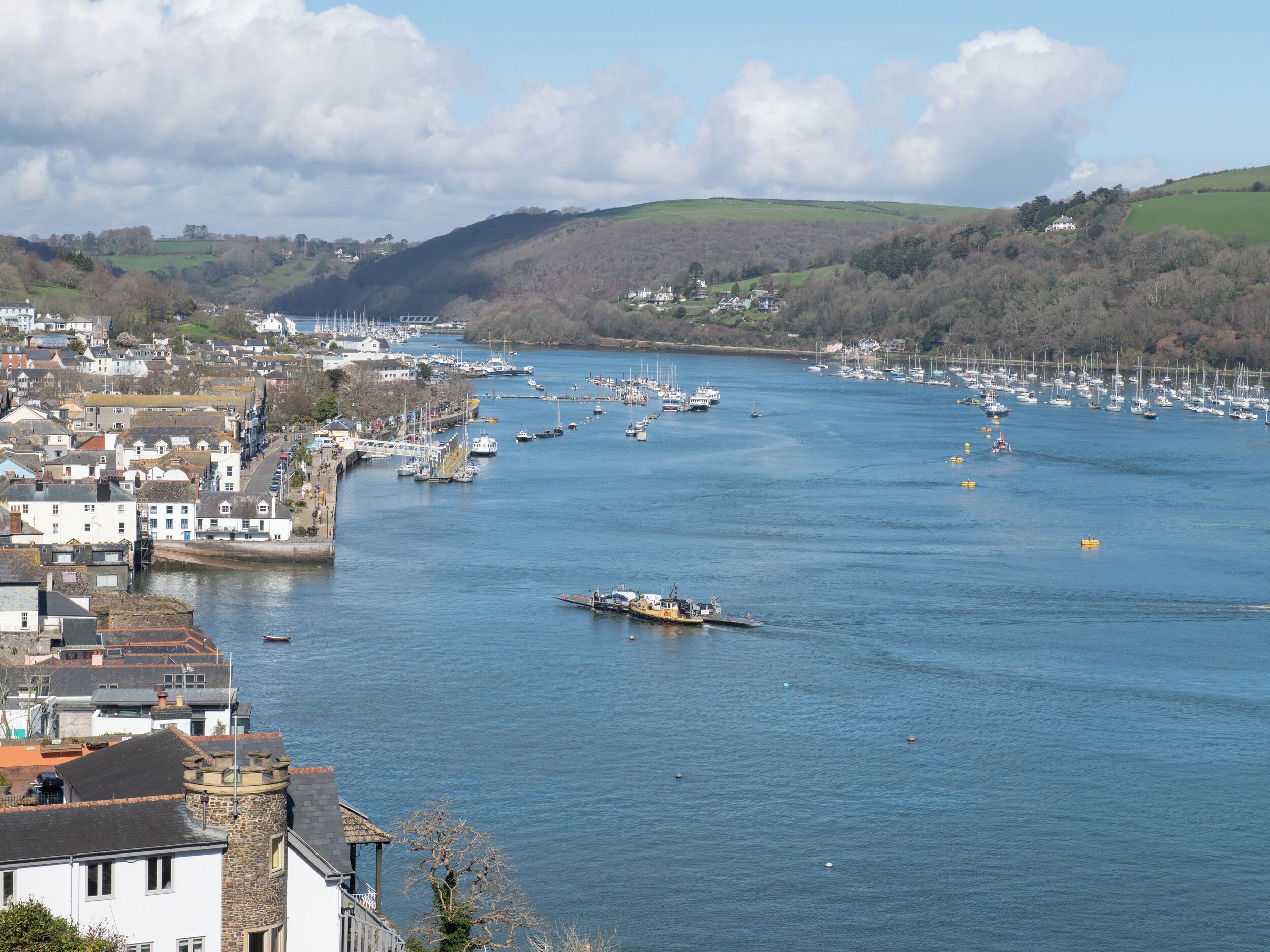 A coastal town with a marina filled with boats, surrounded by hills and residential buildings.