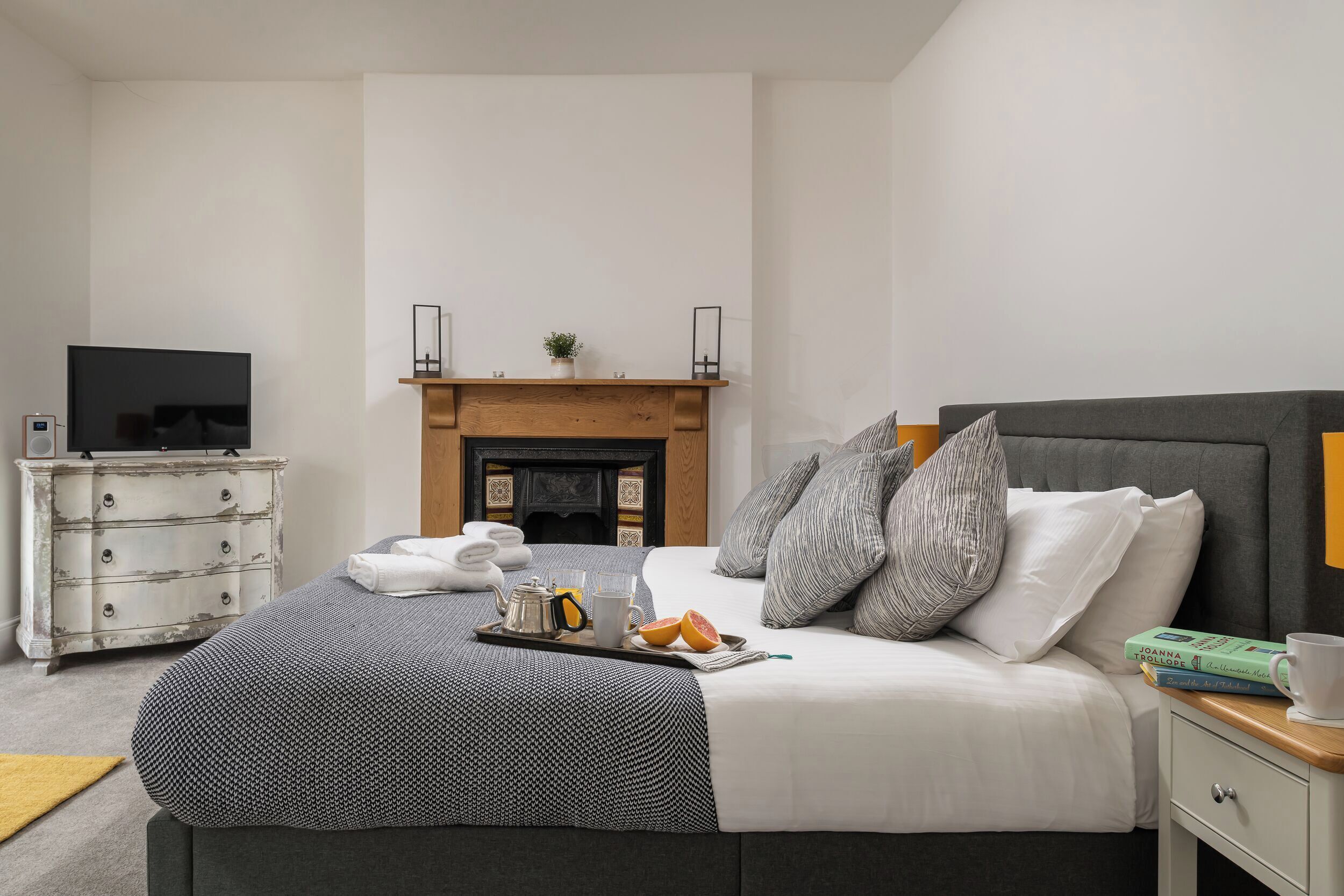 A neatly arranged bedroom with a bed, bedside table, a flat-screen TV, a dresser, and a fireplace.