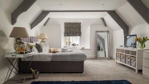Une chambre avec un plafond en pente, un lit avec des oreillers et un livre, une table de chevet avec une lampe, une télévision et une fenêtre avec des rideaux.