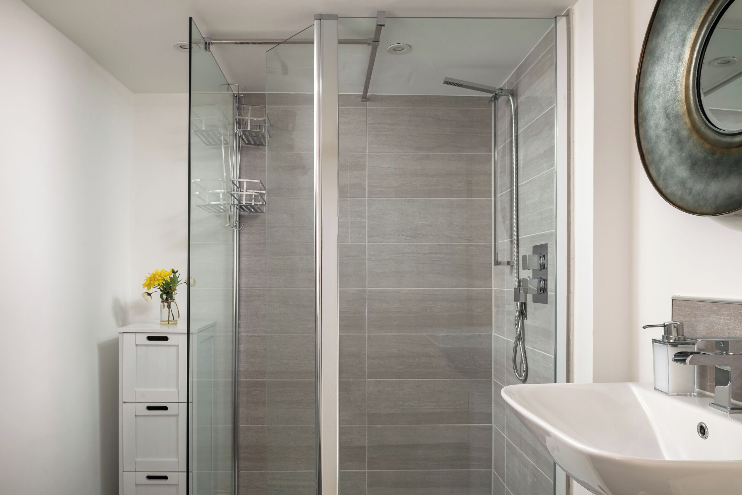 A modern bathroom with a glass-enclosed shower, a white sink, and a vase with flowers on a cabinet.