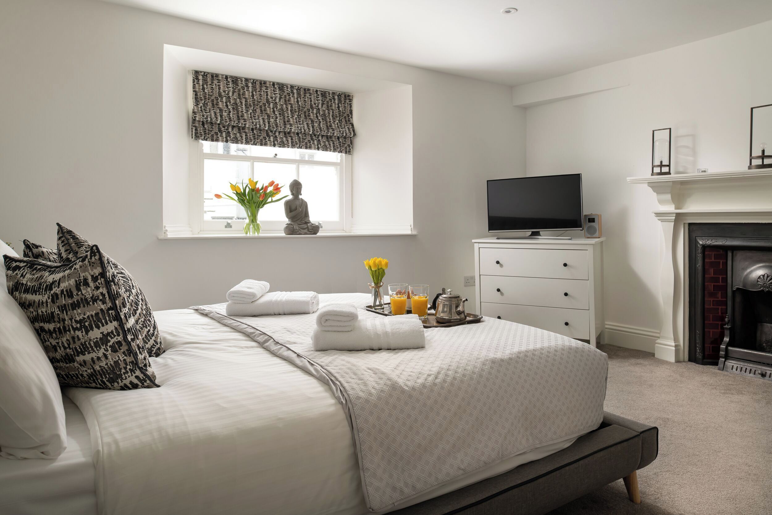 A neatly made bed with a patterned pillow, a bedside table with a flat-screen TV, a vase with flowers, and a fireplace.