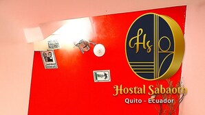 A staircase leading to a room with a red wall, a logo, and a sign indicating 'Hostal Sabaooth Quito - Ecuador.' - HOSTAL SABAOTH (Quito)