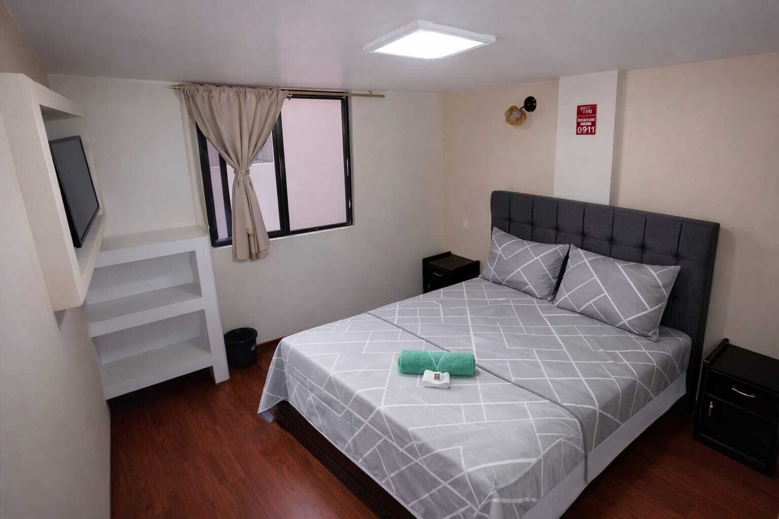 Un dormitorio con cama, televisor montado en la pared, mesita de noche y cortinas en la ventana, además de un detector de incendios.