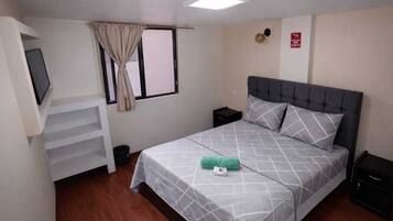 Un dormitorio con cama, televisor montado en la pared, mesita de noche y cortinas en la ventana, además de un detector de incendios.