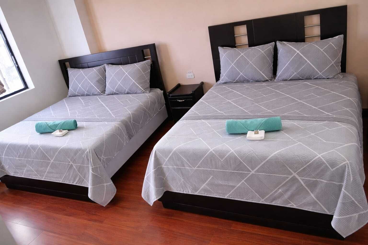 Dos camas individuales con ropa de cama gris y toallas color turquesa, cabeceros de madera y piso de madera.