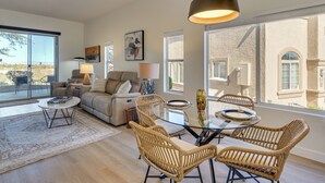 A modern living room with a glass coffee table, wicker chairs, a sofa, and a large window offering a view of a building.