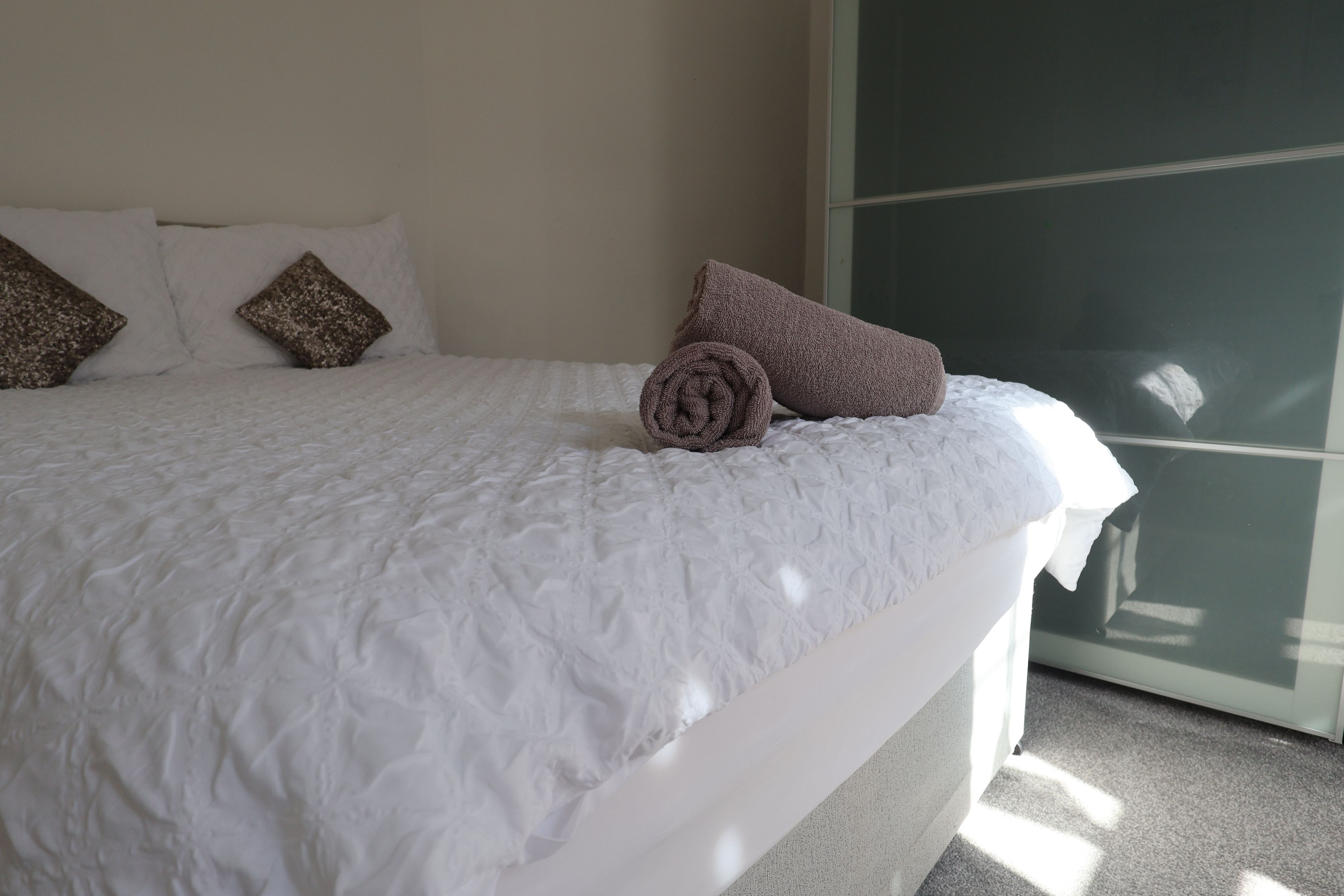 Un lit recouvert d’une couverture blanche à motifs, une serviette marron enroulée et deux coussins décoratifs.