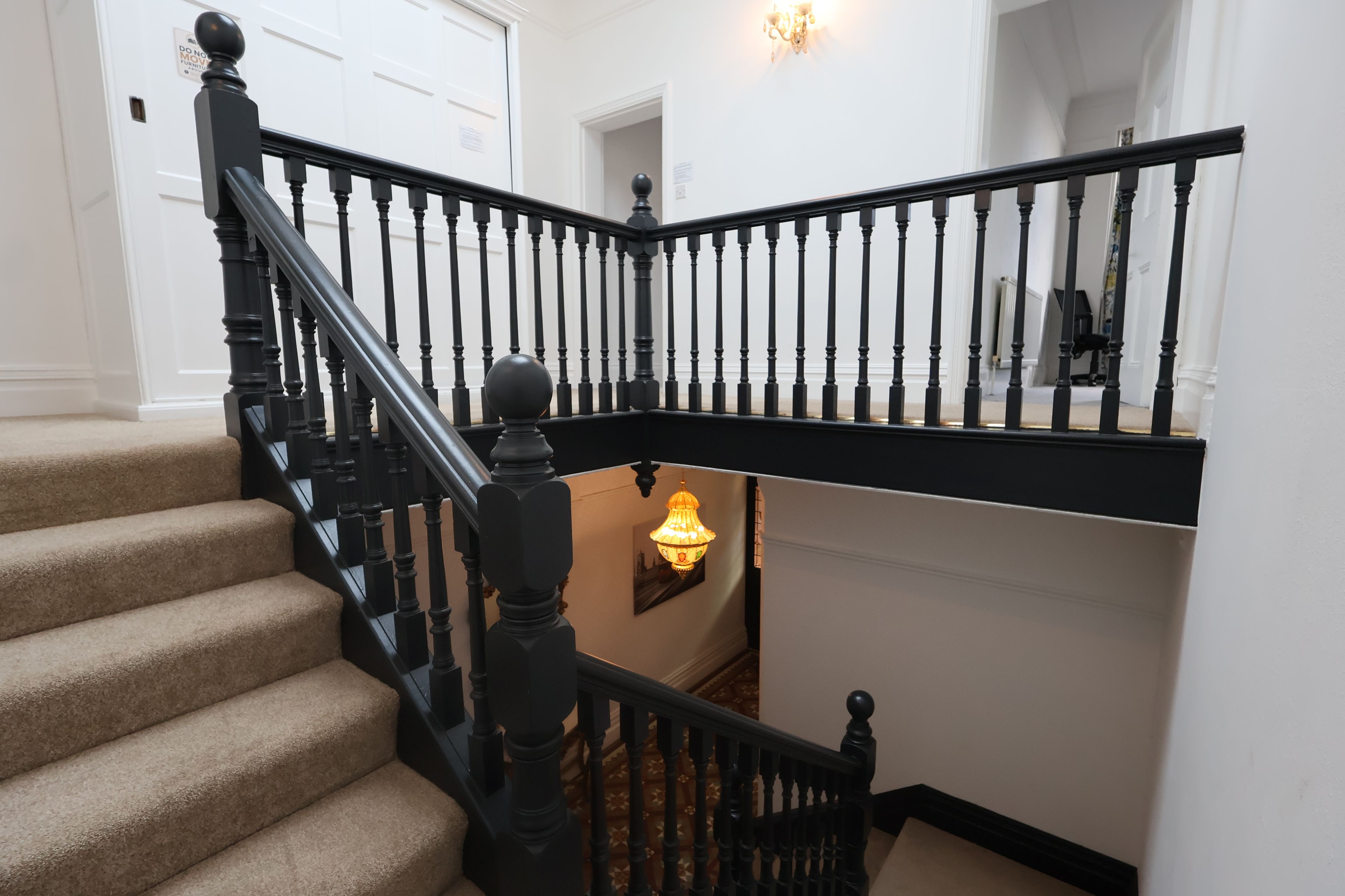 Un escalier avec des balustrades noires et une main courante, des marches recouvertes de moquette et un luminaire fixé au mur.