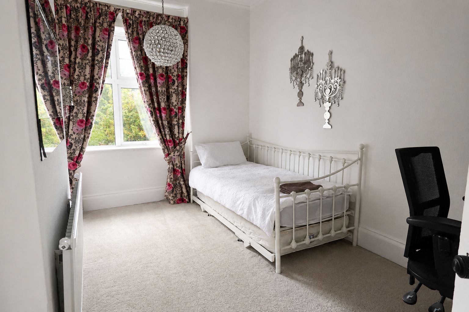 A bedroom with a white metal bed frame, a window with floral curtains, and a chandelier.