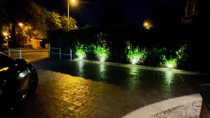 Cena noturna de rua com um carro estacionado à esquerda, postes de luz e arbustos iluminados ao longo da calçada.