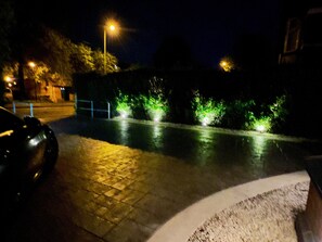 A nighttime street scene with a car parked on the left, streetlights, and illuminated bushes along the sidewalk.