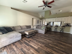 A spacious living room with a large sectional sofa, a coffee table, and a kitchen area with bar stools and wooden flooring.