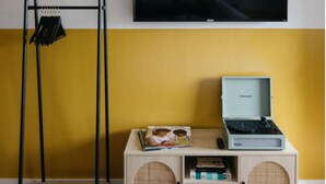 Una habitación con una pared amarilla, un televisor sobre una mesa con un tocadiscos y libros, y una estantería metálica negra con ropa.