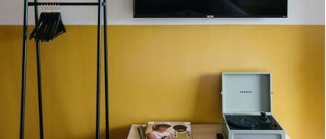 Una habitación con una pared amarilla, un televisor sobre una mesa con un tocadiscos y libros, y una estantería metálica negra con ropa.