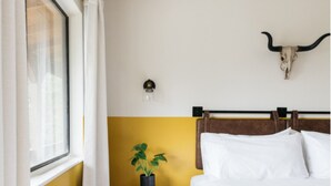 Una habitación con una cama, una ventana con cortinas, una lámpara de pared, una planta sobre una mesita y una decoración con cráneo de vaca.
