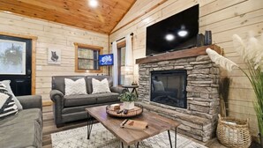 A cozy living room with a stone fireplace, a flat-screen TV, a sofa, and a coffee table with a plant and books. - Cozy Cardinal with Hot Tub (Sevierville)